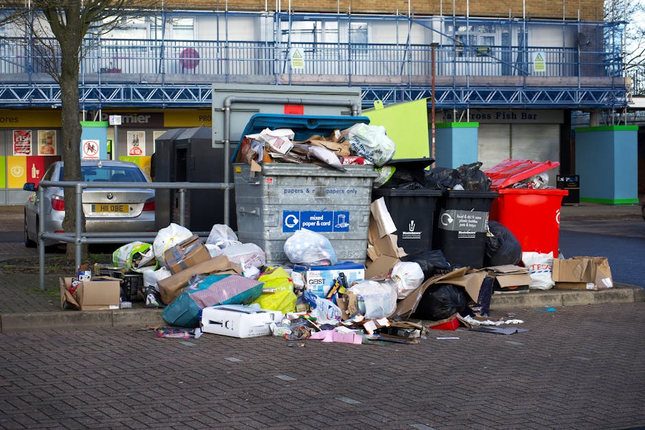 A pile of discarded household rubbish and debris situated on a gravel surface in an outdoor area, with various black and clear plastic bags, some filled with waste, stacked amid a few loose items incl