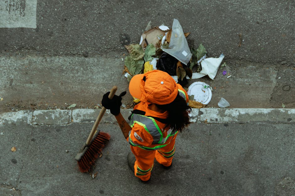 A person dressed in bright orange high-visibility workwear, including a jacket, pants, and a hat, is standing on a paved sidewalk next to a low concrete wall. They are holding a long-handled broom wit