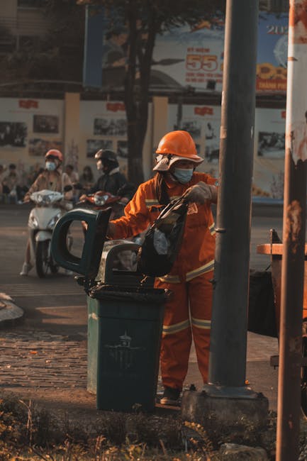 A person dressed in bright orange high-visibility workwear, including a jacket, pants, and a hat, is standing on a paved sidewalk next to a low concrete wall. They are holding a long-handled broom wit
